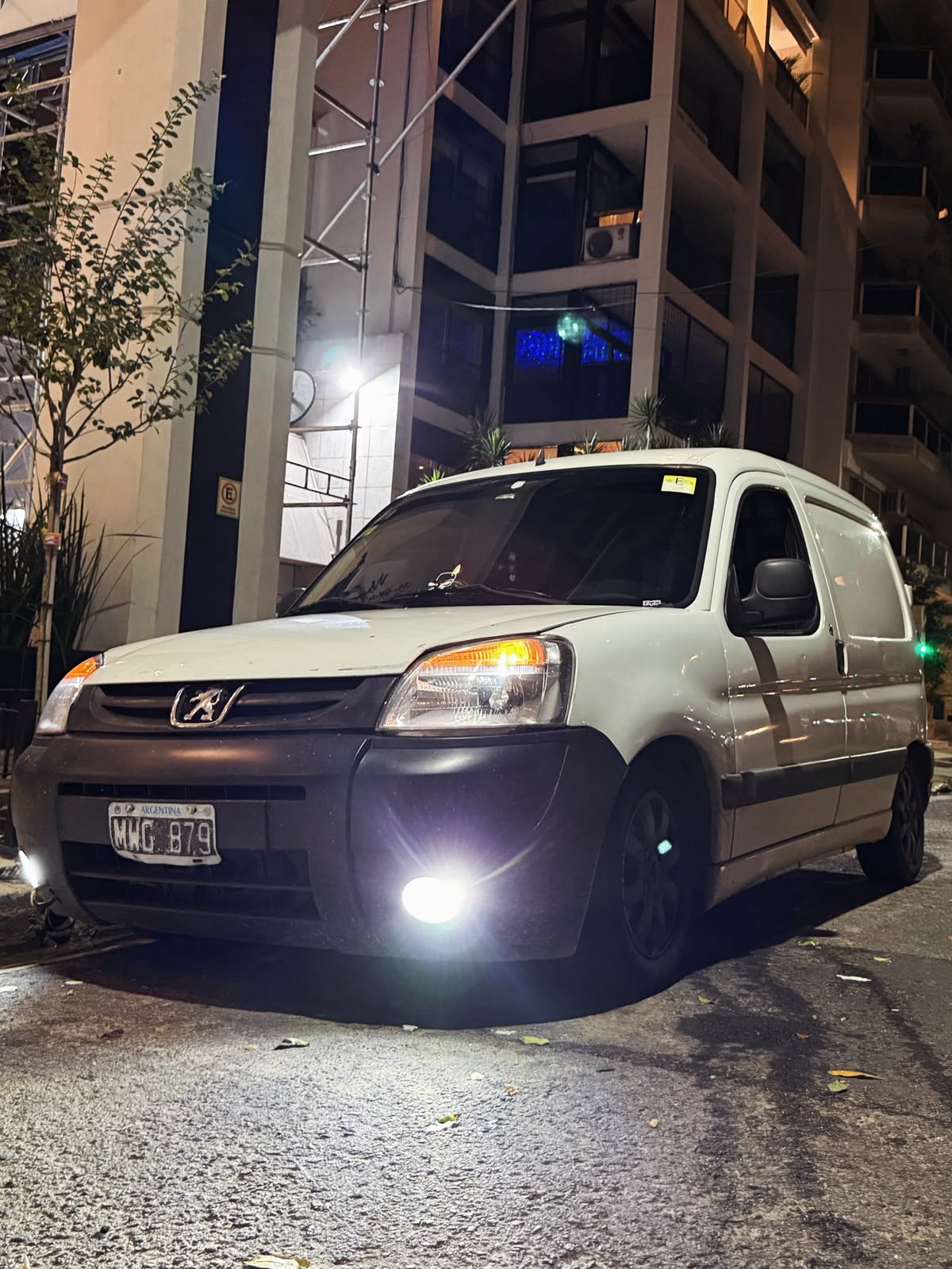 La Peugeot Partner blanca de Facha Fletes, patente argentina, estacionada de noche con las luces encendidas